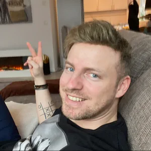 Smiling man on a sofa giving a peace sign, with a tattooed forearm, in a living room featuring a lit fireplace in the background.