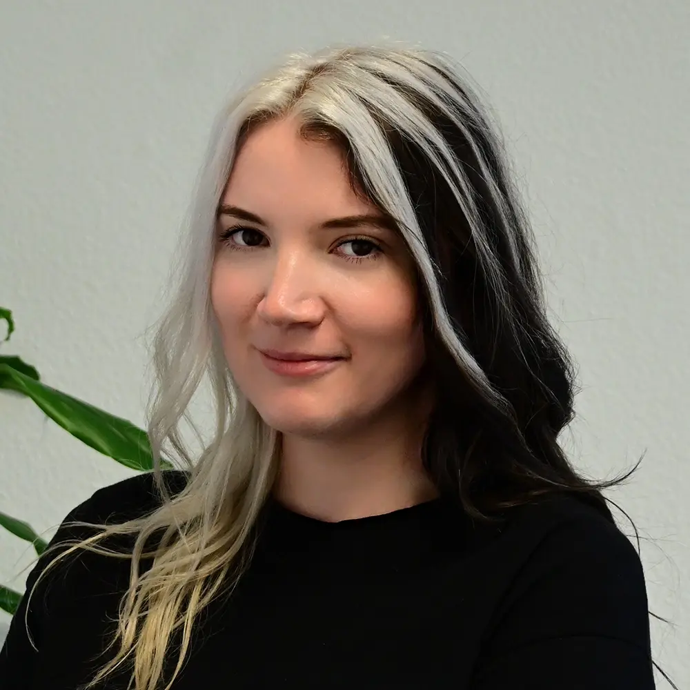 Woman with split black and platinum blonde hair, smiling softly in a black top, neutral background.